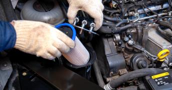 Dłonie w białych rękawiczkach wkładają jasny wkład filtra do pionowej obudowy w silniku wysokoprężnym.