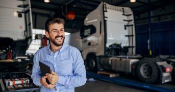 Uśmiechnięty spedytor lub menedżer logistyki trzymający tablet na tle serwisowanych ciągników siodłowych w hali garażowej.
