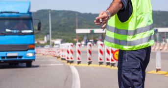 Widok od tyłu na funkcjonariusza służb drogowych w odblaskowej kamizelce i granatowym mundurze, który ręką z lizakiem daje sygnał do zatrzymania nadjeżdżającej niebieskiej ciężarówce. Scena przedstawia rutynową kontrolę ITD na drodze.