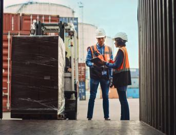 „Pracownicy logistyki w kamizelkach odblaskowych sprawdzają dokumenty przy załadunku palety do kontenera transportowego.