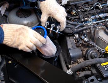 Dłonie w białych rękawiczkach wkładają jasny wkład filtra do pionowej obudowy w silniku wysokoprężnym.