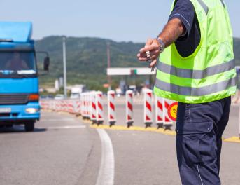 Widok od tyłu na funkcjonariusza służb drogowych w odblaskowej kamizelce i granatowym mundurze, który ręką z lizakiem daje sygnał do zatrzymania nadjeżdżającej niebieskiej ciężarówce. Scena przedstawia rutynową kontrolę ITD na drodze.