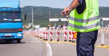 Widok od tyłu na funkcjonariusza służb drogowych w odblaskowej kamizelce i granatowym mundurze, który ręką z lizakiem daje sygnał do zatrzymania nadjeżdżającej niebieskiej ciężarówce. Scena przedstawia rutynową kontrolę ITD na drodze.